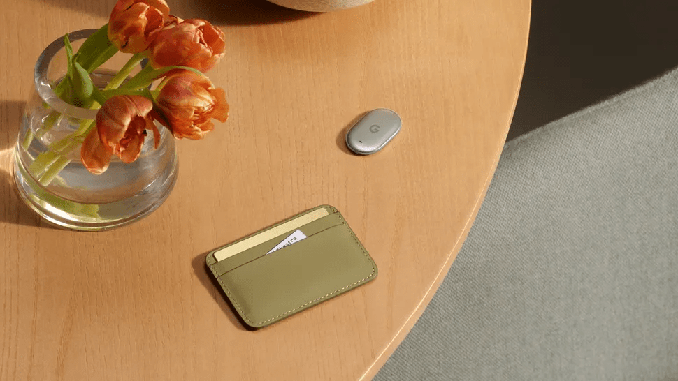 Pixel Watch 5 diventa Health Guardian mentre Google lancia Pixel Tag e nuovi Buds 7 Un piccolo Pixel Tag argentato poggiato su un tavolo di legno accanto a un portafoglio verde oliva e tulipani in vaso.